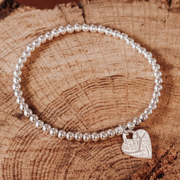 Silbernes Kugelarmband aus 925 Sterling Silber mit Herzanhänger, Vorderseite mit eingraviertem Fingerabdruck, fotografiert auf einer Holzscheibe.