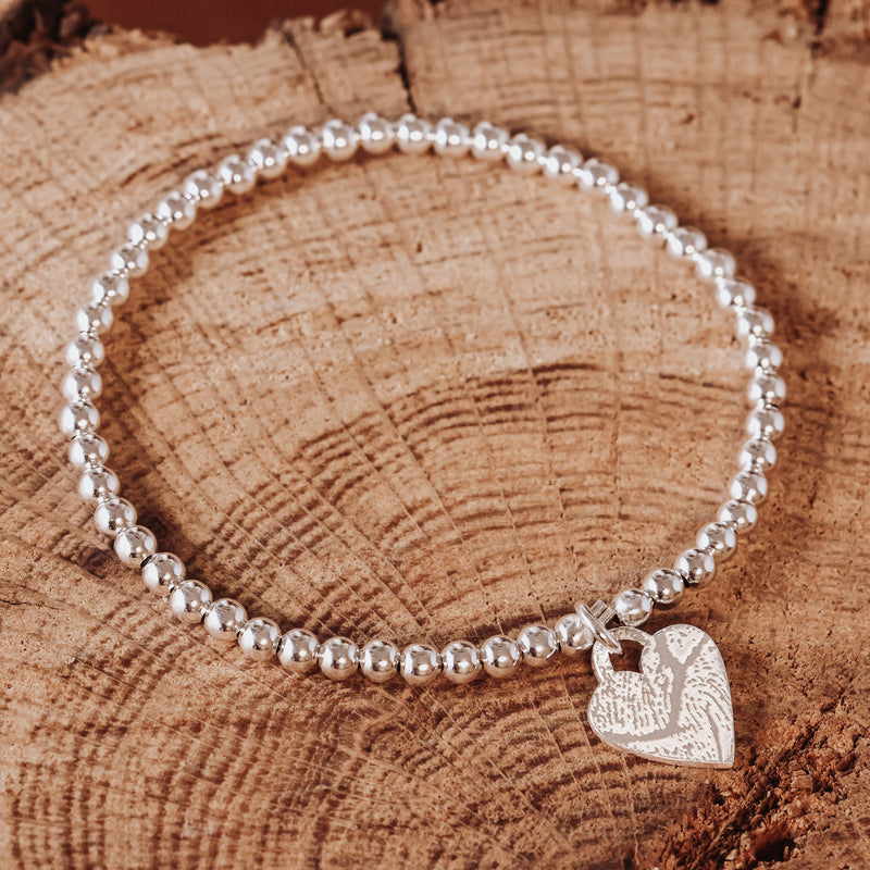 Silbernes Kugelarmband aus 925 Sterling Silber mit Herzanhänger, Vorderseite mit eingraviertem Fingerabdruck, fotografiert auf einer Holzscheibe.