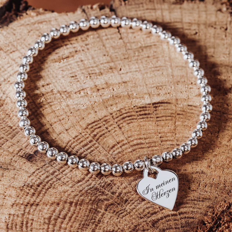 Silbernes 925 Sterling Silber Kugelarmband mit Herzanhänger, Rückseite mit Gravur „In meinen Herzen“, dekorativ auf Holz fotografiert.